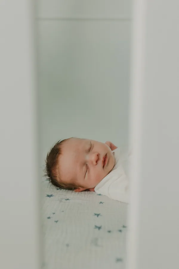 Close-up of a sleeping newborn in a white outfit on a fitted cot sheet. Sleep surface mostly cropped; do not describe this photo as a complete safe-sleep setup.