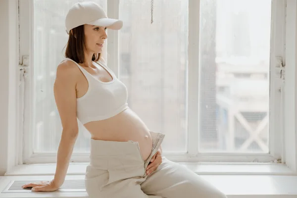 Pregnant woman sitting beside a bright window, resting a hand on her belly.